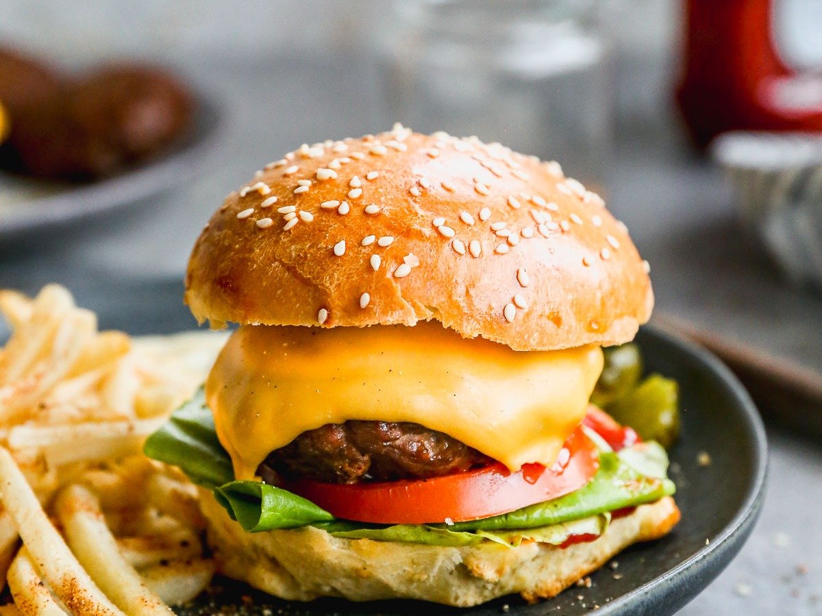 A cheeseburger with lettuce, tomato, and a sesame seed bun on a black plate, accompanied by a side of seasoned fries. A blurred ketchup bottle and two jars are visible in the background on a gray surface.