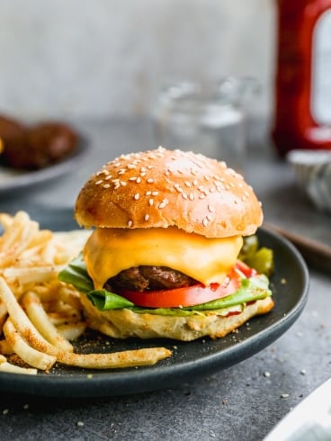 A cheeseburger with lettuce, tomato, and a sesame seed bun on a black plate, accompanied by a side of seasoned fries. A blurred ketchup bottle and two jars are visible in the background on a gray surface.