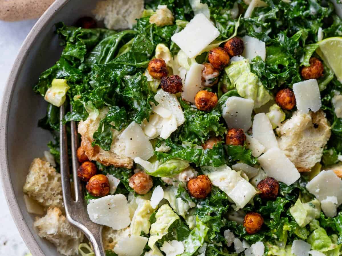 A bowl of kale salad topped with shaved Parmesan cheese, crispy chickpeas, and croutons. A fork rests in the bowl. Lime wedges and a small bowl of dressing are on the side. The background is a light-colored surface.