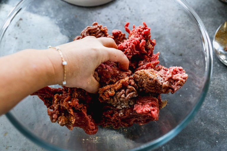 A hand mixes raw ground beef with seasoning in a clear glass bowl on a gray countertop. The meat appears red and is slightly clumped together. The person is wearing a bracelet on the wrist.
