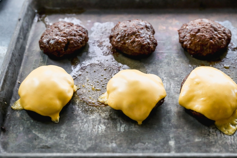 Four hamburger patties on a metal baking sheet; two are topped with melted cheese slices, and two are plain. The cheese is slightly oozing over the sides of the patties.