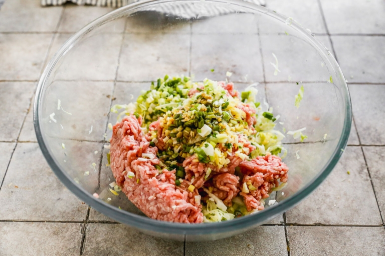 A glass bowl on a tiled countertop containing ground meat mixed with chopped vegetables such as onions and celery.