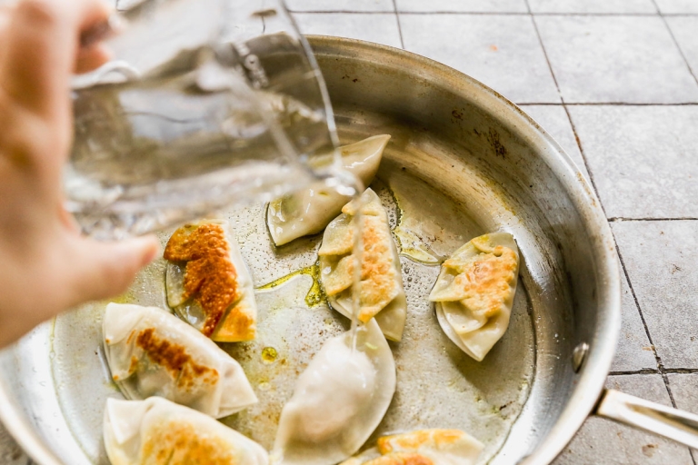 A hand pours water into a pan with sizzling pork dumplings. The dumplings are golden brown on one side, and the pan is set on a tiled surface.