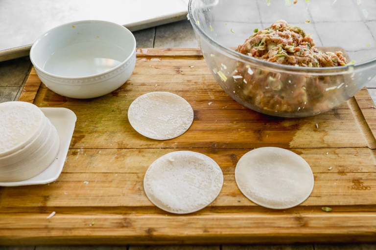 A wooden cutting board with dumpling wrappers, a bowl of meat filling mixture, and a small bowl of water. Dumpling wrappers are laid out, and the meat filling contains visible chopped ingredients.