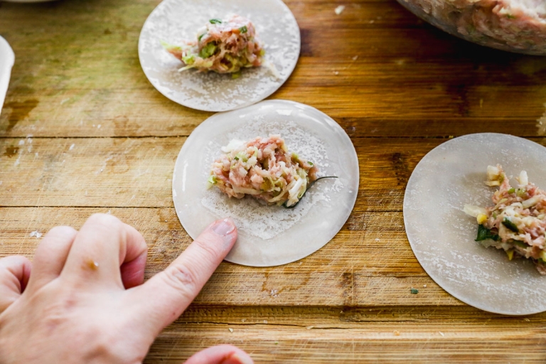 Close-up of a hand assembling dumplings on a wooden surface. Circular dumpling wrappers are filled with a mixture of minced meat and vegetables. One wrapper is being prepared for sealing with a finger pressing along its edge.