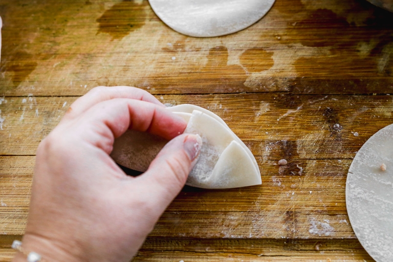 A person's hand folding a dumpling wrapper on a wooden surface. The round dough is partially filled and being pleated. Other wrappers are visible in the background.