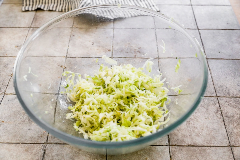 A clear glass bowl on a tiled surface contains finely shredded cabbage, not dressed. A striped cloth is partially visible in the background.