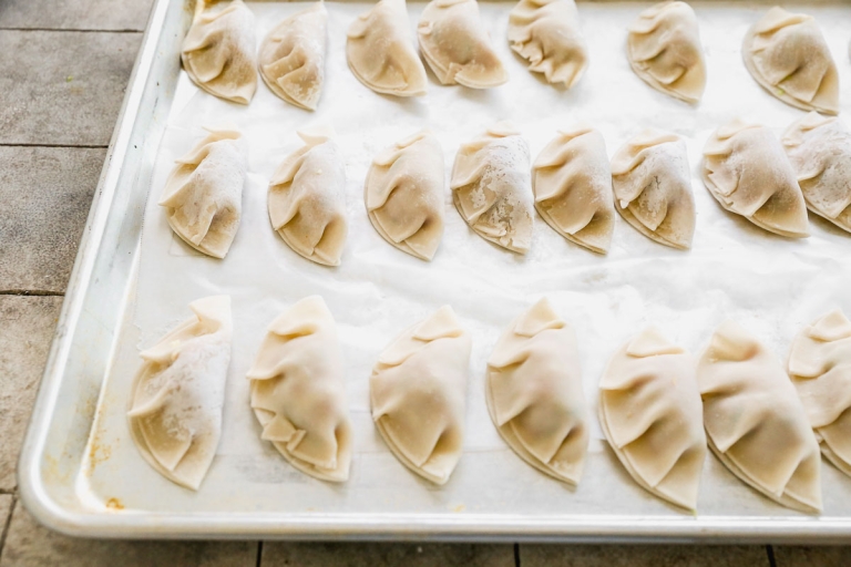 A baking tray filled with neatly arranged, uncooked dumplings, each half-moon shaped, on a lightly floured surface. The dumplings appear homemade, with pleated edges and are ready for cooking.