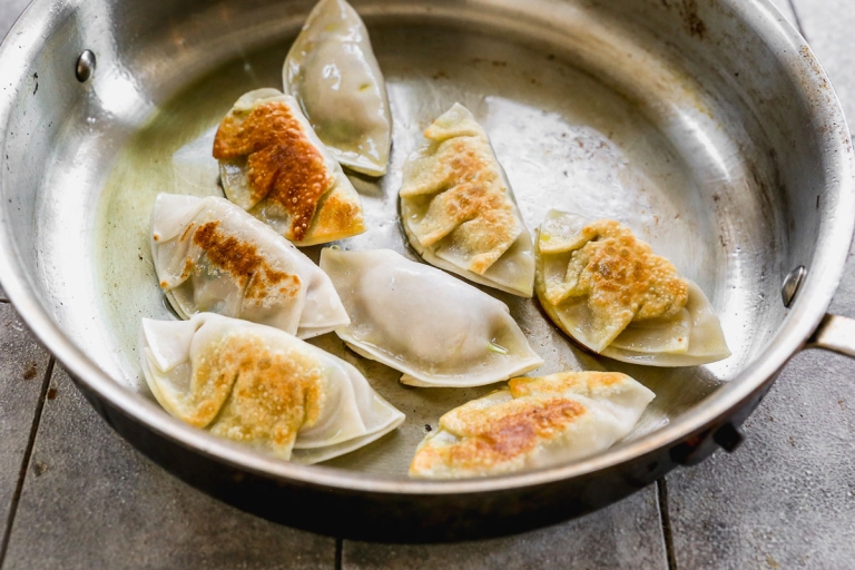 A stainless steel pan containing several pan-fried dumplings, some with golden brown crispy edges, rests on a wood surface. The dumplings are arranged randomly in the pan, displaying a mix of colors from light beige to golden brown.
