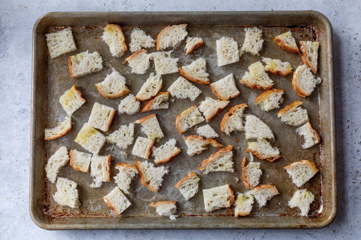 A baking sheet holds an even layer of small, rustic pieces of bread, coated with oil, ready to be toasted. The bread is arranged randomly on the metal tray, which has a slightly worn appearance.