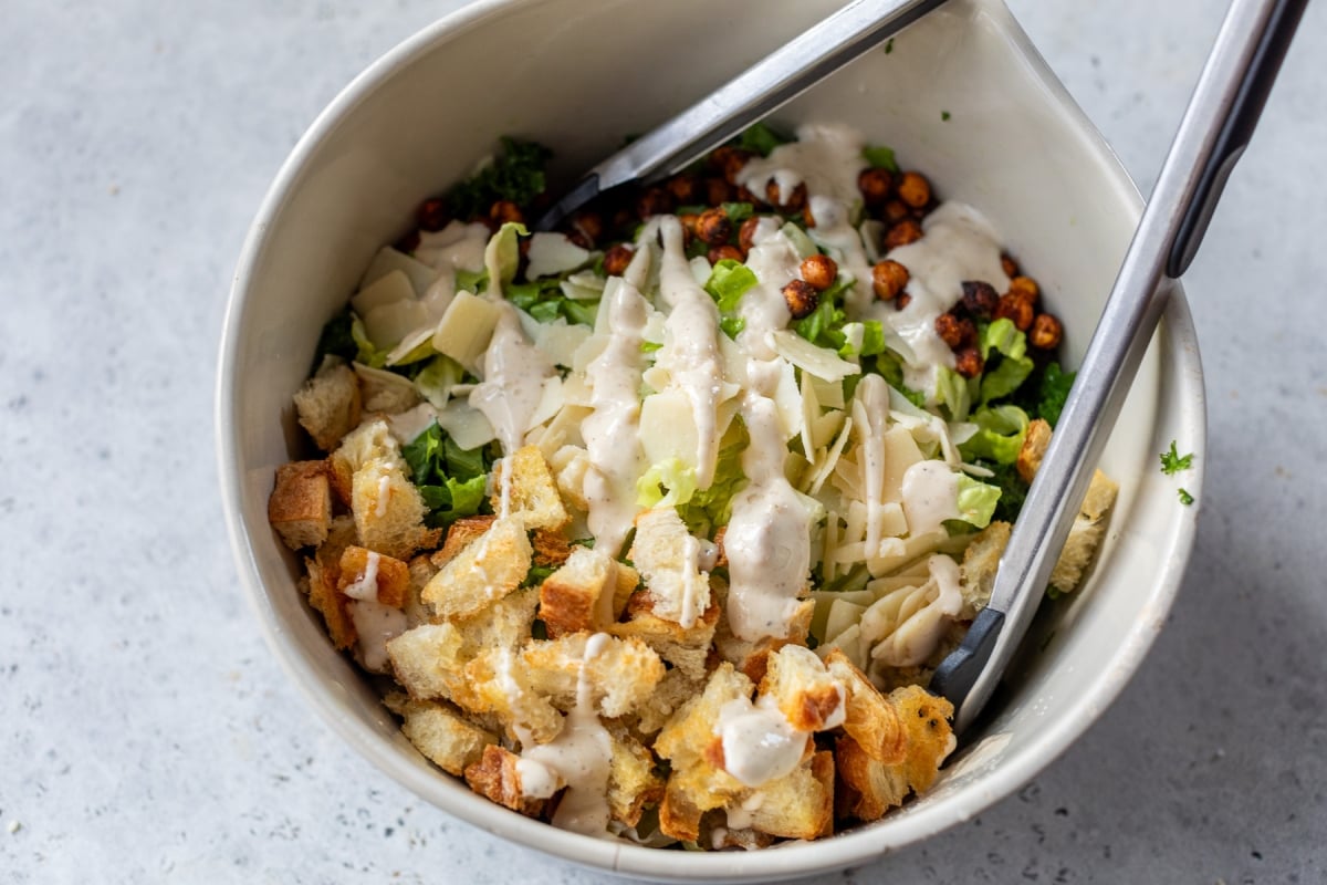A bowl of Caesar salad featuring chopped lettuce, croutons, shaved Parmesan cheese, crispy roasted chickpeas, and creamy dressing. Tongs are placed in the bowl, and the background is a light gray surface.