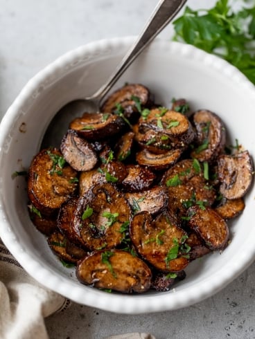 A white bowl filled with sliced air fryer mushrooms garnished with chopped parsley. A spoon rests inside the bowl, and fresh parsley is visible in the background. The surface is light gray, creating a contrast with the dish.