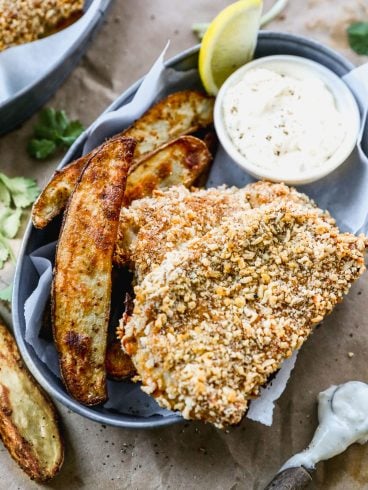 A plate with crispy breaded fish fillet, thick-cut potato wedges, and a small cup of creamy dipping sauce garnished with a lemon wedge. The surface beneath appears to be rustic brown paper with herbs scattered around.
