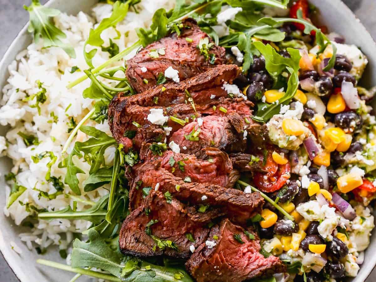 A bowl filled with white rice, topped with slices of grilled steak, arugula, and a mix of black beans, corn, red onion, and peppers. A small bowl of sliced red onions is in the background. The dish is garnished with herbs.