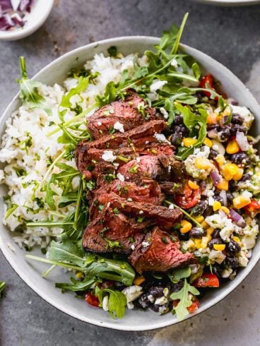 A bowl filled with white rice, topped with slices of grilled steak, arugula, and a mix of black beans, corn, red onion, and peppers. A small bowl of sliced red onions is in the background. The dish is garnished with herbs.