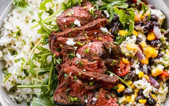A bowl filled with white rice, topped with slices of grilled steak, arugula, and a mix of black beans, corn, red onion, and peppers. A small bowl of sliced red onions is in the background. The dish is garnished with herbs.