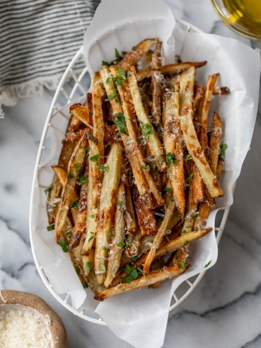 A white basket with parchment paper holds a serving of golden, crispy truffle fries garnished with chopped parsley. The background features a striped cloth, a small bowl of grated cheese, and a glass of oil on a marble surface.