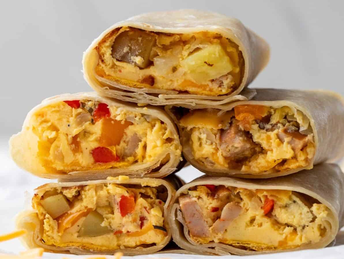 Stack of breakfast burritos cut in half to show fillings, including scrambled eggs, diced potatoes, red peppers, and sausage, with visible cheese. They are arranged on a white surface with scattered cheese shreds nearby.