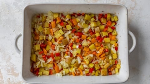 A baking dish filled with diced yellow potatoes, red and yellow bell peppers, and onions. The vegetables are seasoned and ready to be roasted, displayed on a light-colored countertop.