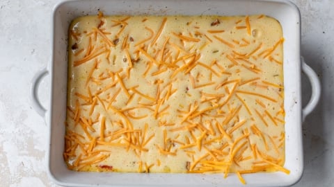 A rectangular baking dish filled with egg mixture, topped with shredded cheddar cheese. The mixture appears to include small visible ingredients like herbs or spices. The dish rests on a lightly textured, light-colored surface.