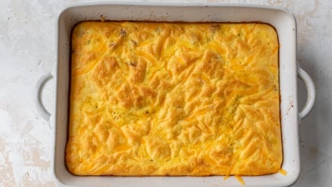A baked egg omelet in a rectangular white casserole dish with handles, featuring a golden-brown, slightly textured surface. The background is a light, textured surface.