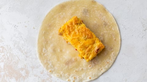 A flour tortilla with a rectangular piece of a yellow egg and cheese omelette placed at the center on a light textured surface.