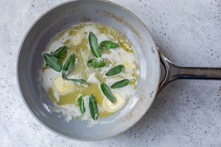 A skillet with melted butter and fresh sage leaves sizzling on a gray countertop.