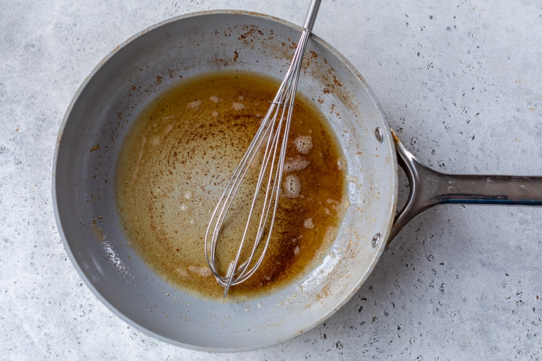 A stainless steel whisk rests in a frying pan with melted butter and brown bits on the bottom. The pan is on a light gray textured surface, suggesting preparation of a sauce or roux.