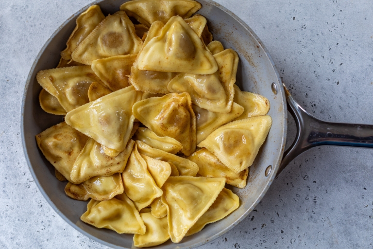A pan filled with cooked butternut squash ravioli pieces, lightly browned, on a textured gray surface. The pasta has a golden hue and is arranged in overlapping layers within the pan.