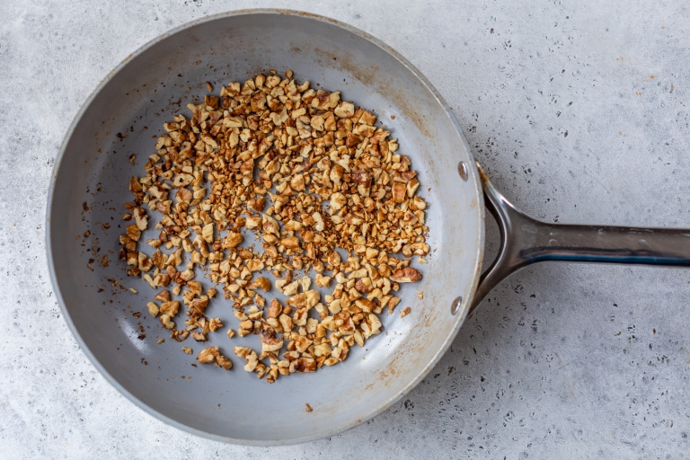 Chopped nuts are being toasted in a gray frying pan set on a textured light gray surface. The nuts are golden brown, indicating they are nearly done. The pan has a long metal handle.