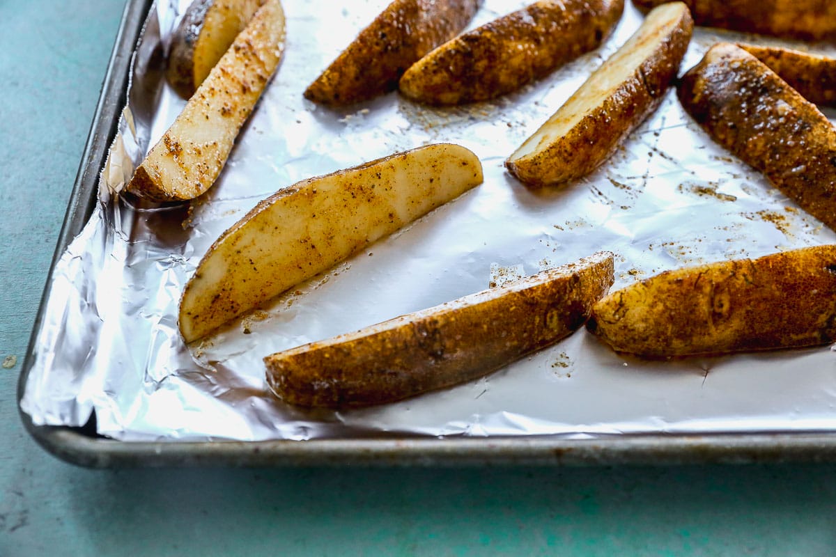 Seasoned potato wedges are arranged on a foil-lined baking sheet. The wedges have a golden-brown, seasoned coating, ready for baking. The background is slightly out of focus, emphasizing the potatoes.