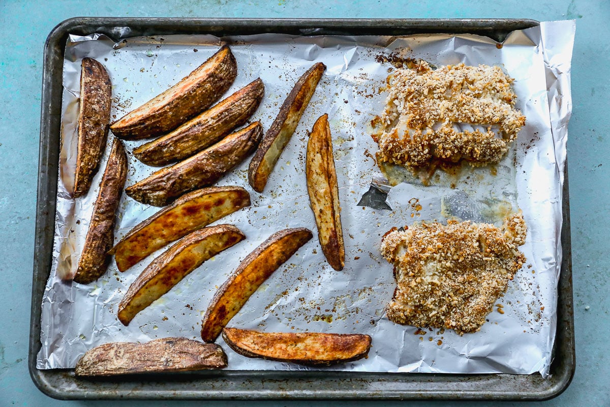 A baking sheet lined with foil holds several seasoned potato wedges on the left and two breaded fish cutlets on the right, both baked and lightly browned. The sheet is set against a light blue background.