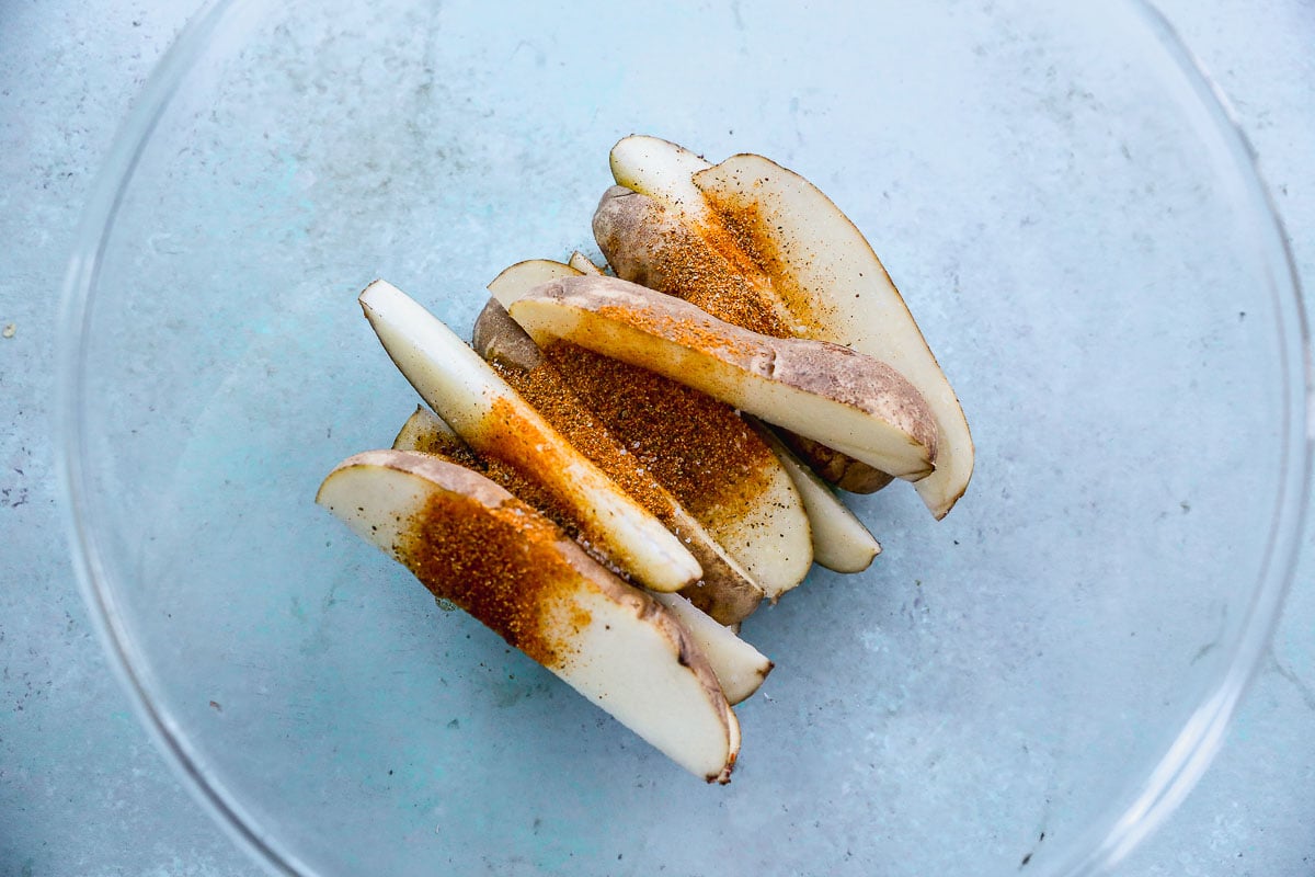 Slices of raw potato in a clear bowl are seasoned with a reddish-brown spice blend, against a light blue background.