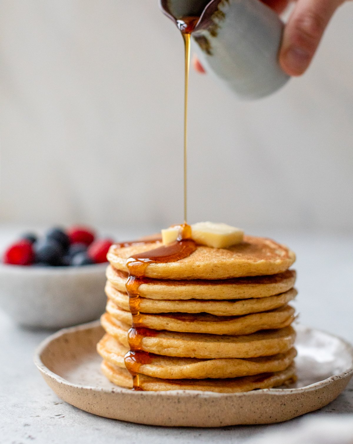 A hand pours syrup over a tall stack of cottage cheese pancakes topped with a pat of butter.