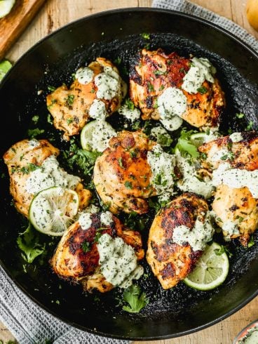 Cilantro lime chicken thighs garnished with fresh cilantro and lime slices, served in a black skillet and topped with a creamy green sauce. The dish sits on a wooden surface with a textured gray cloth underneath.