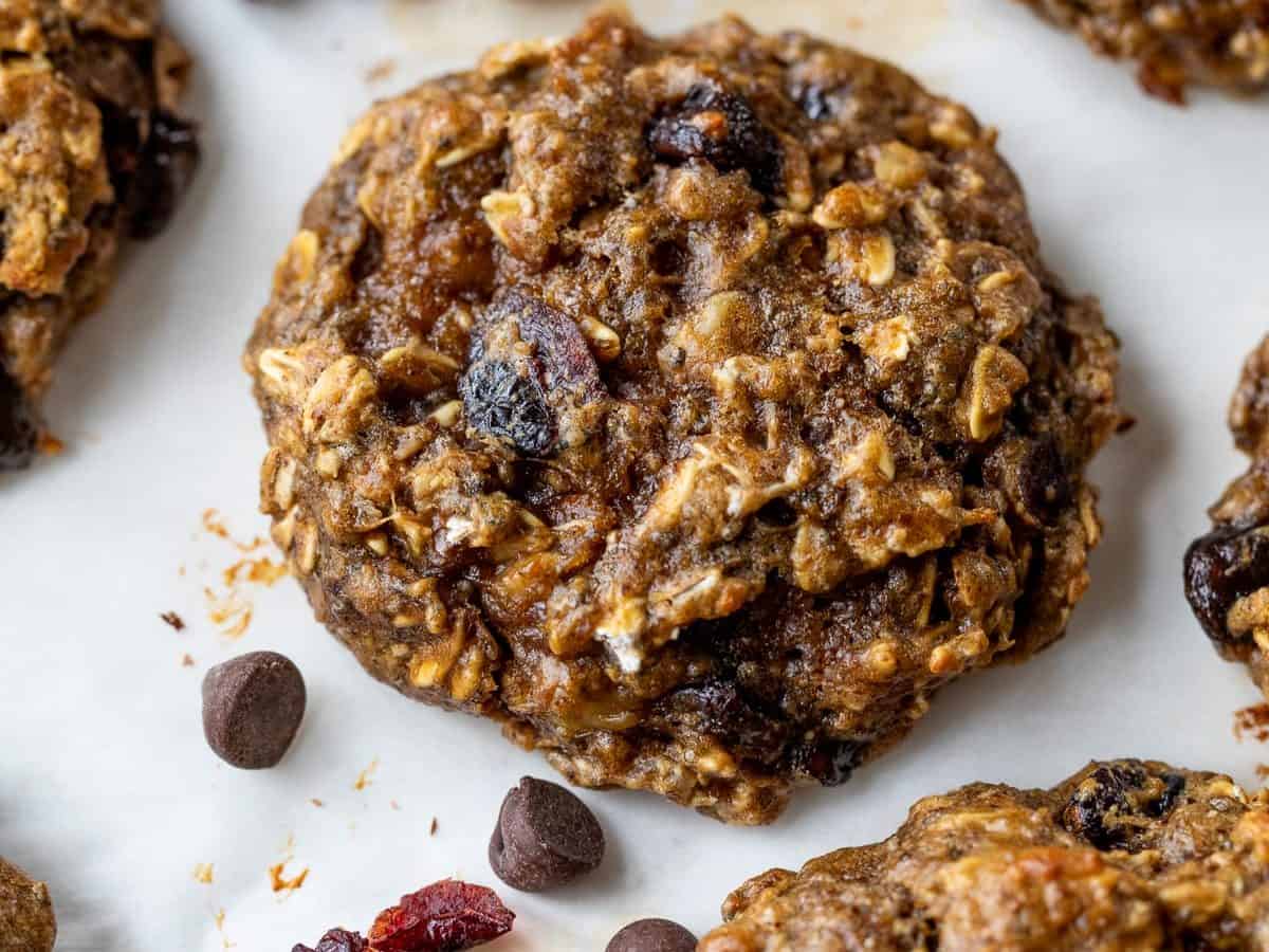 A close-up of oatmeal protein cookies with chocolate chips and dried cranberries on a white surface, with a few chocolate chips and cranberries scattered nearby.
