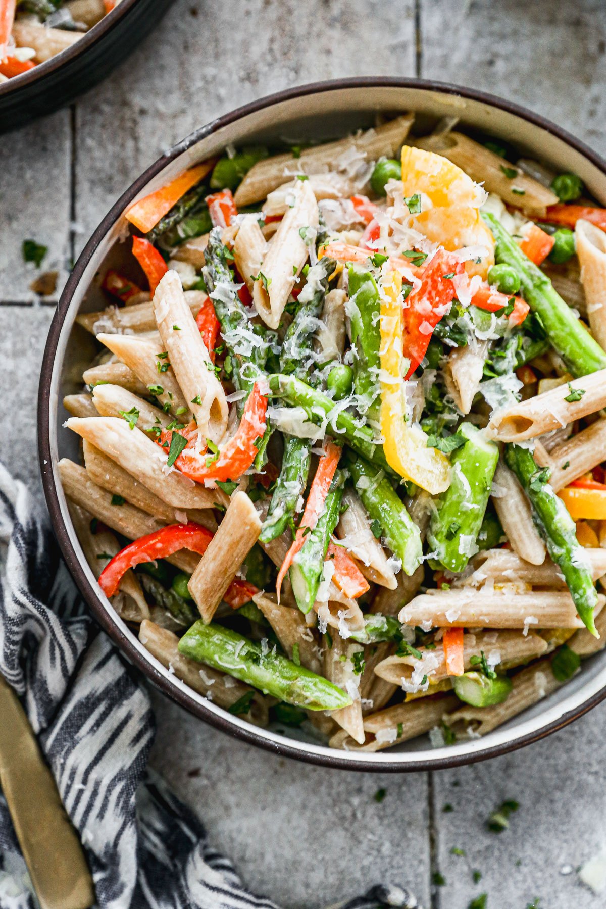 A bowl of pasta primavera with colorful bell peppers, asparagus, and grated cheese, garnished with chopped herbs. The dish is set on a rustic surface with a patterned napkin nearby.