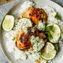 A plate of white rice topped with two pieces of cilantro lime chicken, drizzled with creamy herb sauce and garnished with fresh lime slices and cilantro. The plate sits on a textured gray cloth.