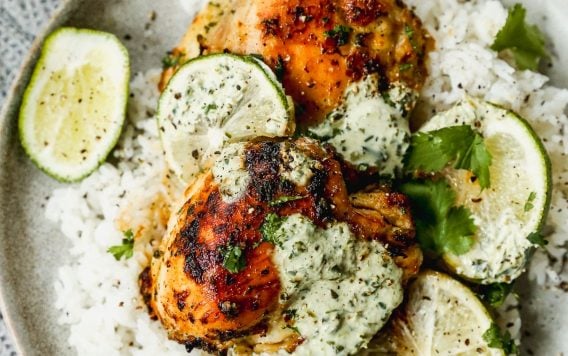 A plate of white rice topped with two pieces of cilantro lime chicken, drizzled with creamy herb sauce and garnished with fresh lime slices and cilantro. The plate sits on a textured gray cloth.
