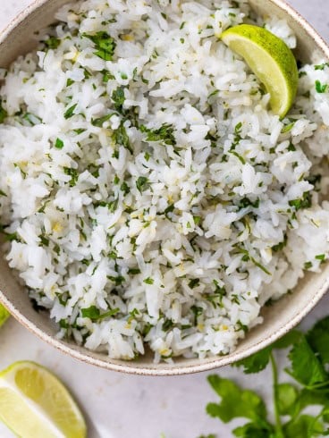 A bowl of cilantro lime rice, garnished with a wedge of lime. Extra lime wedges and fresh cilantro are on the table nearby.
