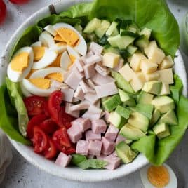 A bowl of chef salad with lettuce, sliced boiled eggs, diced tomatoes, cubed cheese, sliced cucumbers, diced ham, and avocado chunks. Whole tomatoes and an avocado are nearby. A striped cloth is partially visible at the bottom.