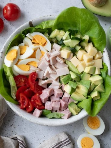 A bowl of chef salad with lettuce, sliced boiled eggs, diced tomatoes, cubed cheese, sliced cucumbers, diced ham, and avocado chunks. Whole tomatoes and an avocado are nearby. A striped cloth is partially visible at the bottom.