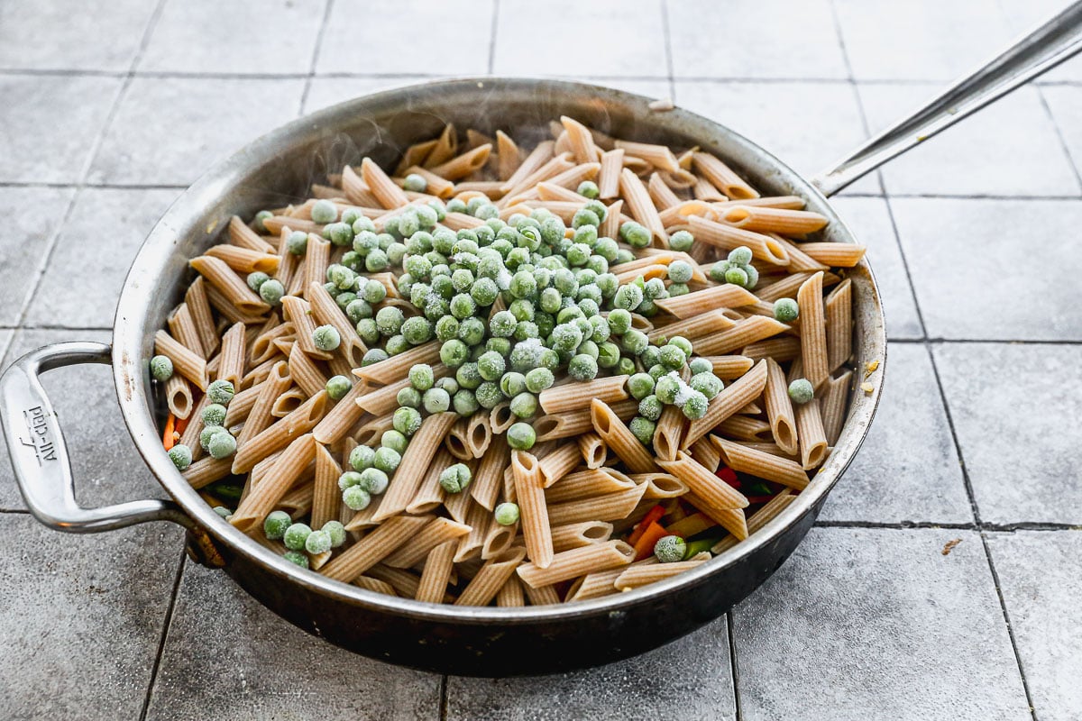 A large pan filled with uncooked whole wheat penne pasta and a pile of frozen green peas sits on a tiled countertop.