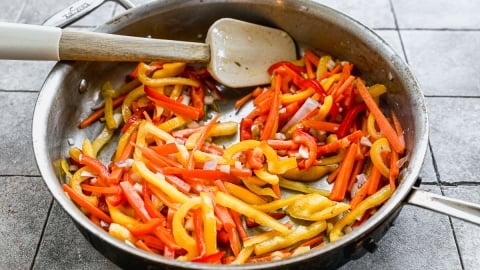 A skillet filled with sautéed strips of red, yellow, and orange bell peppers and onions, with a white spatula resting inside, on a tiled surface.