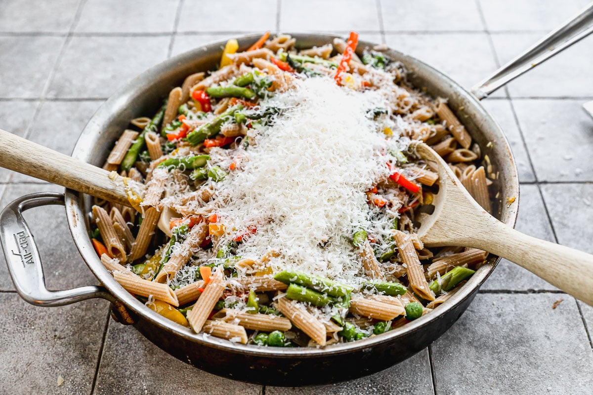 A skillet filled with penne pasta, green beans, and sliced red bell peppers, topped with a generous mound of grated cheese; two wooden spoons rest on either side of the pan.