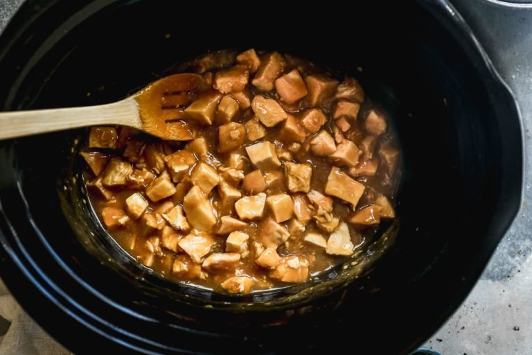 Orange chicken cooking in a black slow cooker, with a wooden spoon resting inside. Steam is rising from the mixture, indicating it is hot and freshly prepared.
