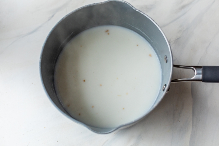 A small pot containing milk, with some small floating steel cut oats, is sitting on a light marble countertop. Steam is rising from the liquid, indicating it is hot. The pot has a black handle.
