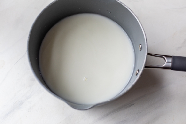 A pot containing warm milk sits on a light surface. The milk appears smooth, creating a simple and clean composition. The pot has a black handle.