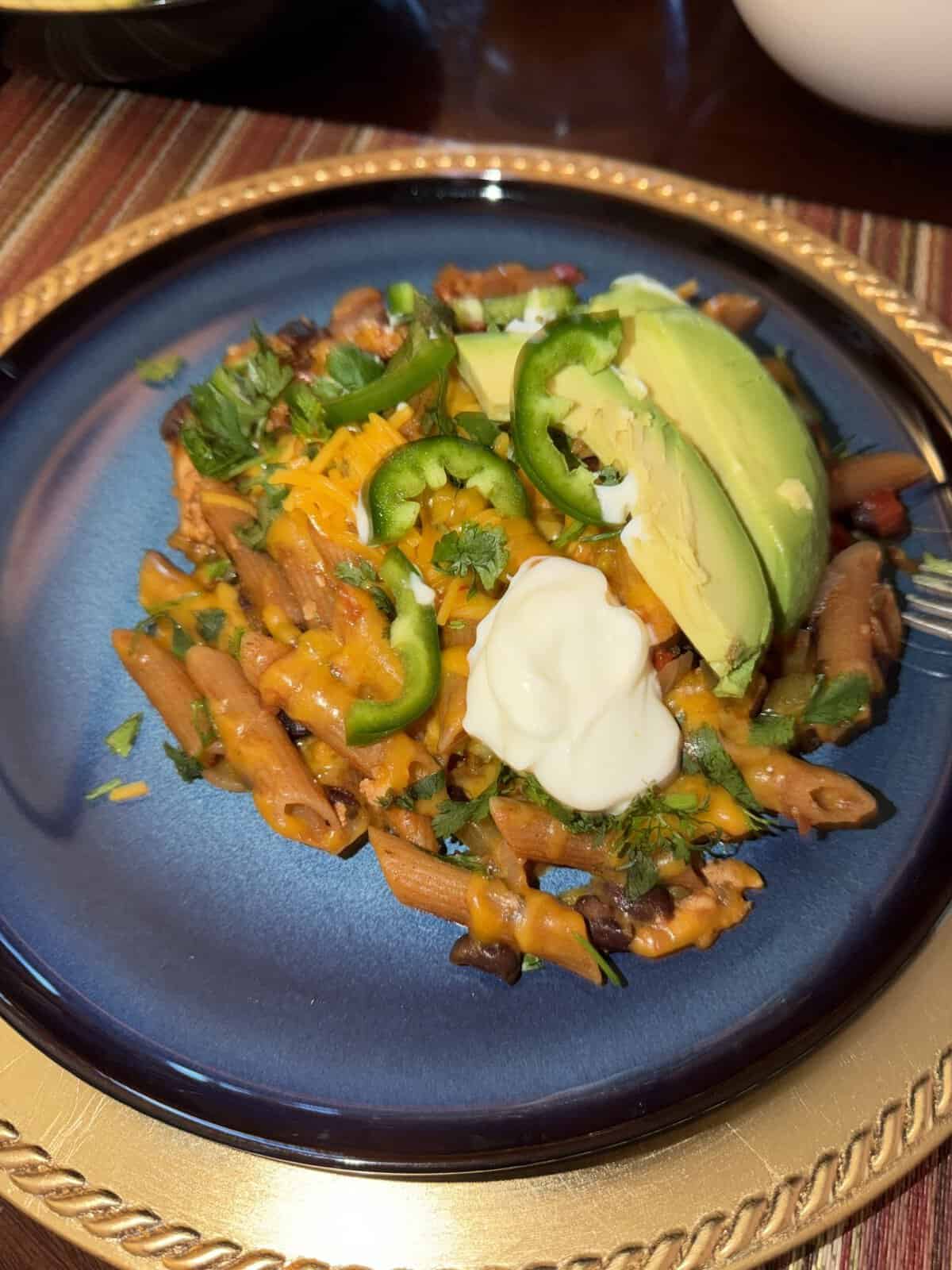 A serving of cheesy pasta topped with avocado slices, jalapeño, sour cream, and herbs on a blue plate.