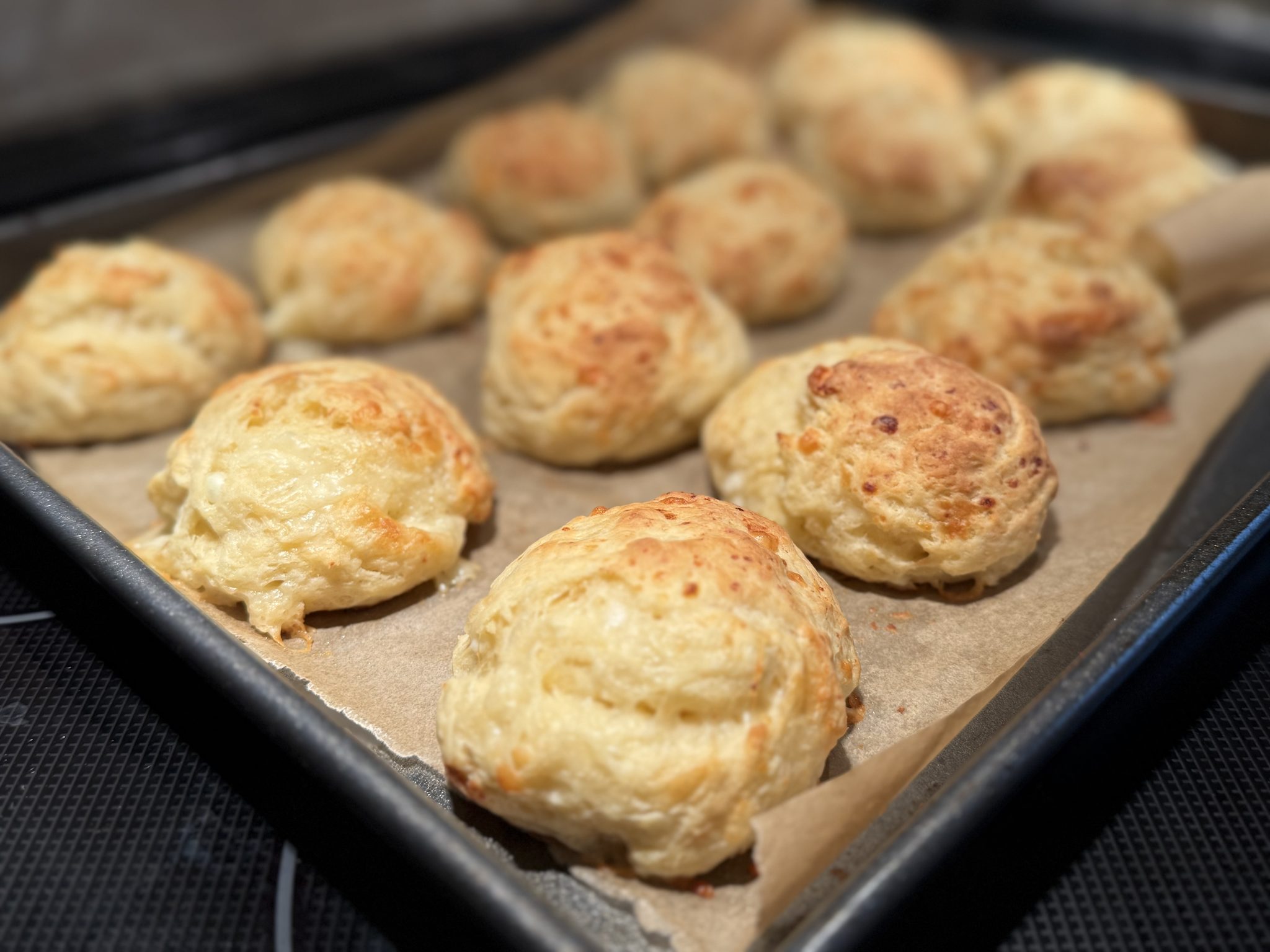 A baking sheet lined with parchment paper holds several golden-brown, freshly baked cheese scones. The scones are fluffy and slightly uneven in shape, with a light, crispy exterior.